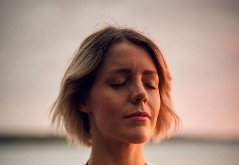 Headshot of a woman with chin-length blond hair, eyes closed, looking serene, with a burred out sunset behind her.