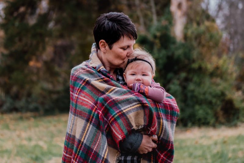 Photo of a woman with short dark hair standing and holding a baby that's looking toward the camera, with a plaid blanket wrapped around them. The woman is kissing the baby's head, and the baby's fist is in its mouth.