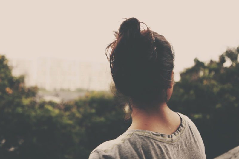 Self-compassion. Image of young woman with long dark hair in a messy bun, looking away from the camera towards some trees in the distance.