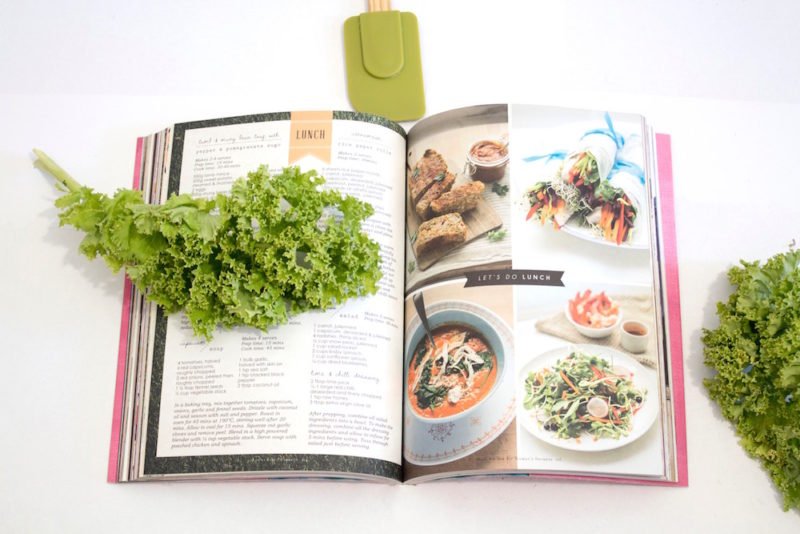 Myths about registered dietitians: Image of an open cookbook surrounded by leaves of curly kale and a green rubber spatula.