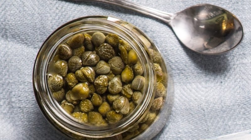 Photo of a jar of capers and a small spoon on a light blue linen cloth