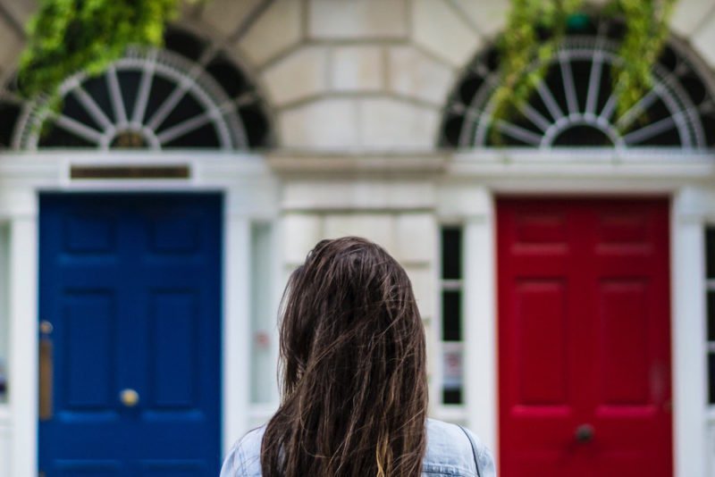 Default behaviors: View of the back of a woman with long, dark hair as she looks at two identical doors, one blue, one red, in the side of a elegant stone building with ornate semi-circular windows above each door.