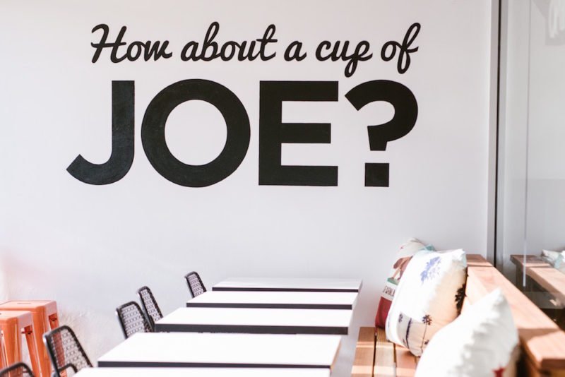 Default behaviors. Photo of a wall in a coffee shot, with "How about a cup of JOE?" painted in black letters on the white wall. The word Joe is very large in a plain, all caps font; the rest of the words are smaller, in a cursive font.