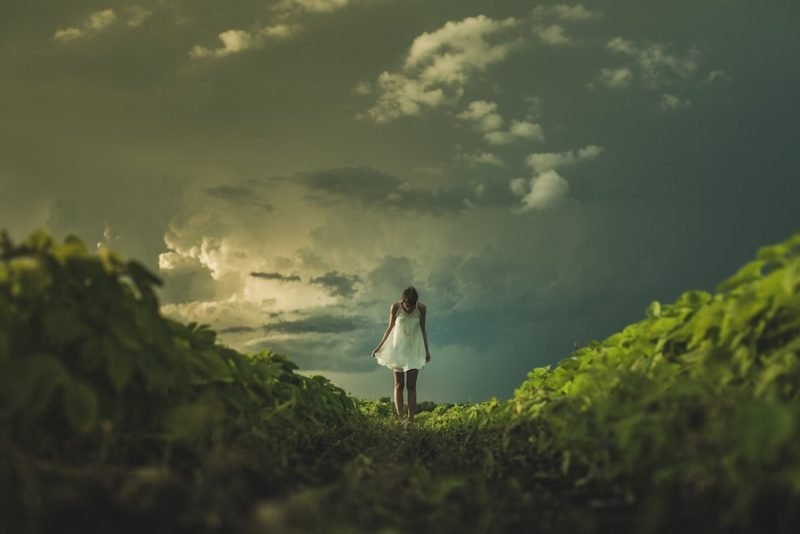 Perfectionism eating disorders. Photo of young woman in a floaty, thigh-length sleeveless dress, staring at her feet as she stands on a plant-covered ridge, with dark, stormy skies behind her.