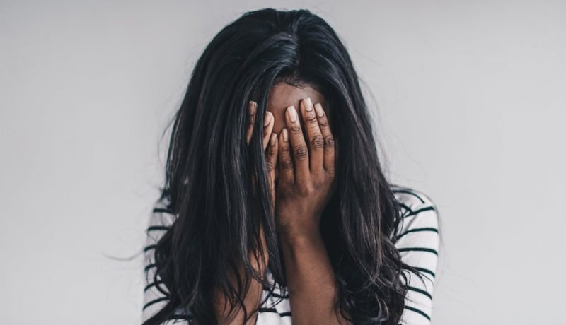 Perfectionism eating disorders. Photo of a young woman with long, dark hair and dark skin, wearing a white T-shirt with black stripes. Her hands are over her face, as if she is crying or otherwise in despair.
