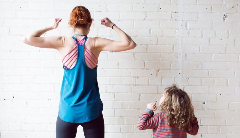 Beauty myth. Photo of a young woman in athletic wear flexing her biceps, while a young child with long hair watches her.