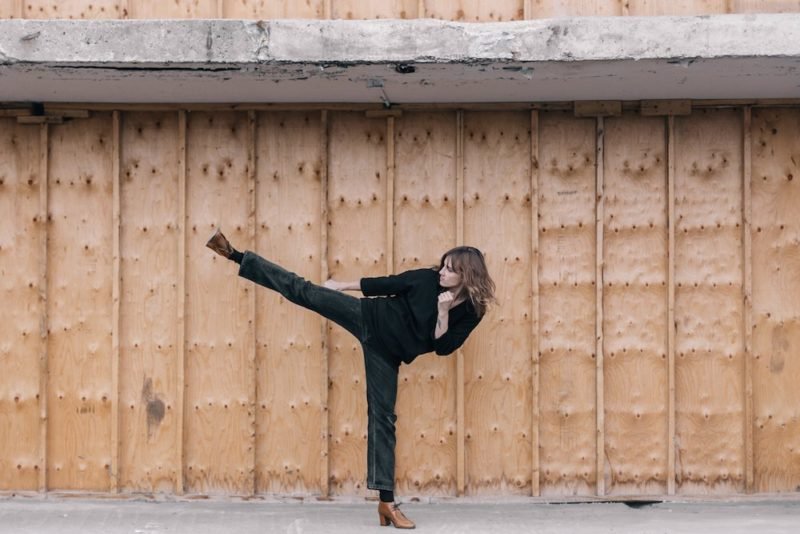 Beauty myth. Photo of young woman in black jeans, a black long-sleeved knit shirt, and brown high-heeled ankle boots doing a high karate kick in front of a industrial-looking wall with covered in plywood