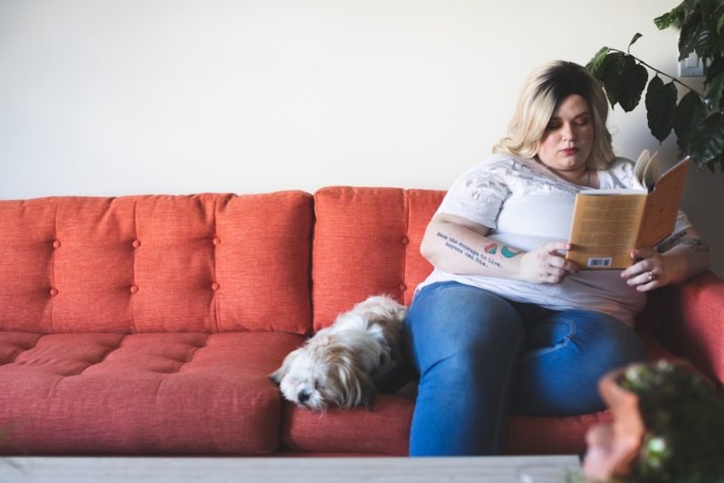 Weight stigma reading list: A young blonde woman in a larger body sitting on an orange couch reading a book, with a small fluffy white dog laying next to her.