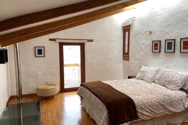 Minimalism: Photo of a bedroom with rustic white walls, a sloped ceiling with wood beams, and simple furnishings (a bed, a wicker ottoman, and some small paintings on the walls)