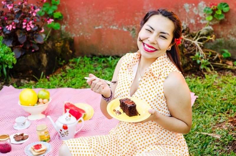 Intuitive eating pleasure can include enjoying a piece of chocolate cake, guilt free, as this woman in a sleeveless white-and-yellow polka dot dress is doing in this photo, smiling brightly with dark pink lipstick, sitting on a pink picnic blanket set with a tea pot and tea cups.