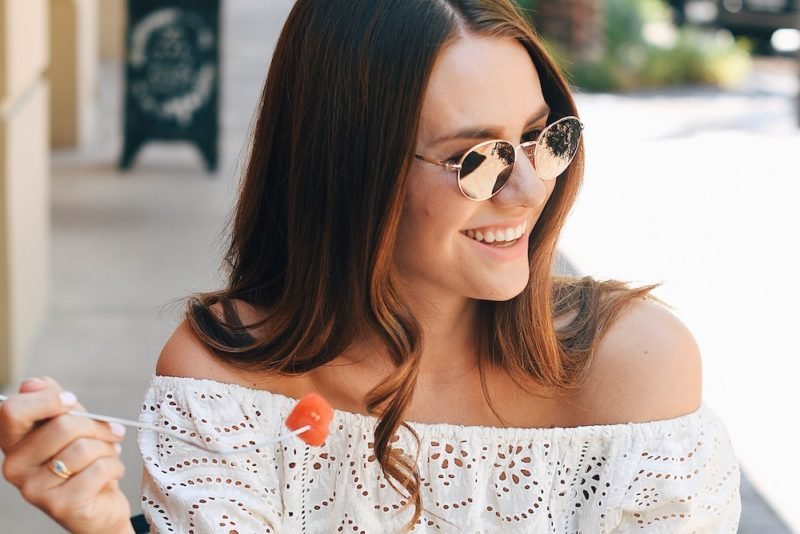 Photo of a young woman with long brown hair, mirrored sunglasses and a white, eyelet off-the-shoulder blouse, holding a fork with a piece of watermelon on it, smiling as she looks to her left.