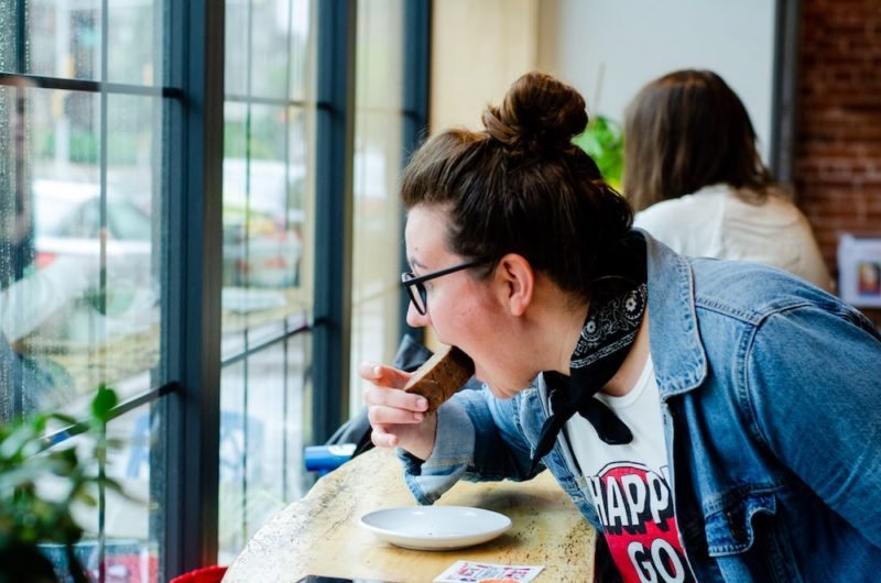 Photo of a young woman with her hair in a bun, wearing a blue denim shirt over a graphic T-shirt eating a slice of banana bread while looking out the window at a coffee shop.