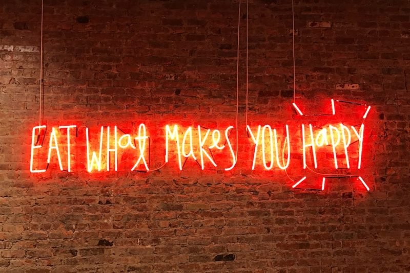 Intuitive eating pleasure is partly about eating what makes you happy. This photo is of a red neon sign that says "Eat What Makes You Happy" in front of a brick wall.