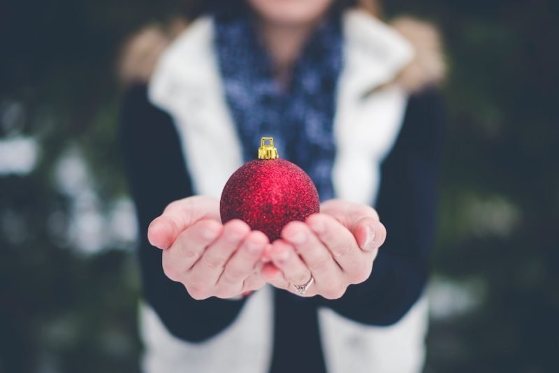 Photo of a woman in a fleece vest and a scarf holding out a red glittery Christmas tree ornament towards the camera.