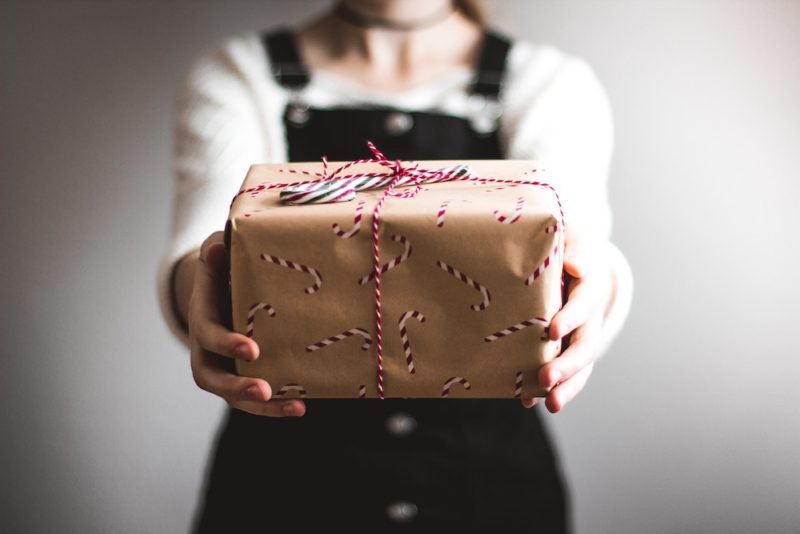 What do you plan to gift yourself for 2020. What would make your year better. Photo of a young woman in a white shirt and dark overalls, holding out a wrapped present towards the camera.