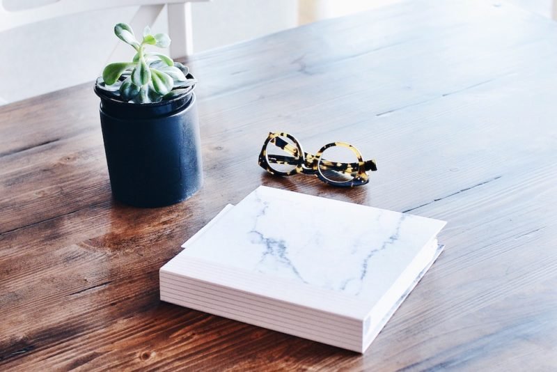 Gift yourself with space to yourself this year. Photo of a book with a white and gray faux marble cover, a pair or tortoiseshell eyeglasses, and a small succulent plant in a black pot on a brown lacquered plank table top.