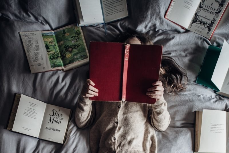 Gift yourself with activities you enjoy, such as reading. Photo of young girl laying on her back on a bed with a gray comforter, surrounded with open books, her face hidden by a book she's holding above her head.