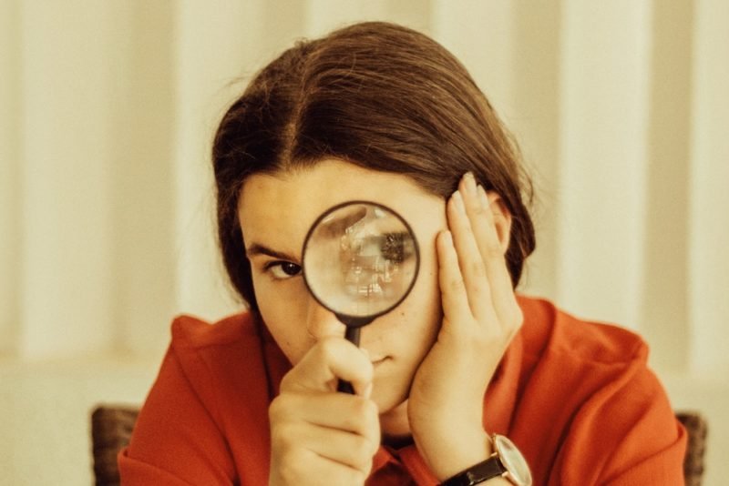 Part of honoring hunger is being curious when you're hungrier than usual. Photo of a young woman with brown hair and a red blouse, peering through a magnifying glass.