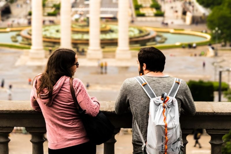 Part of honoring hunger means asserting your needs with others, if needed. Photo of a young woman in a pink hoodie and a young man in a gray long-sleeved shirt and a light blue backpack, leaning against an ornate concrete railing with some ornate columns and fountains in the distance.