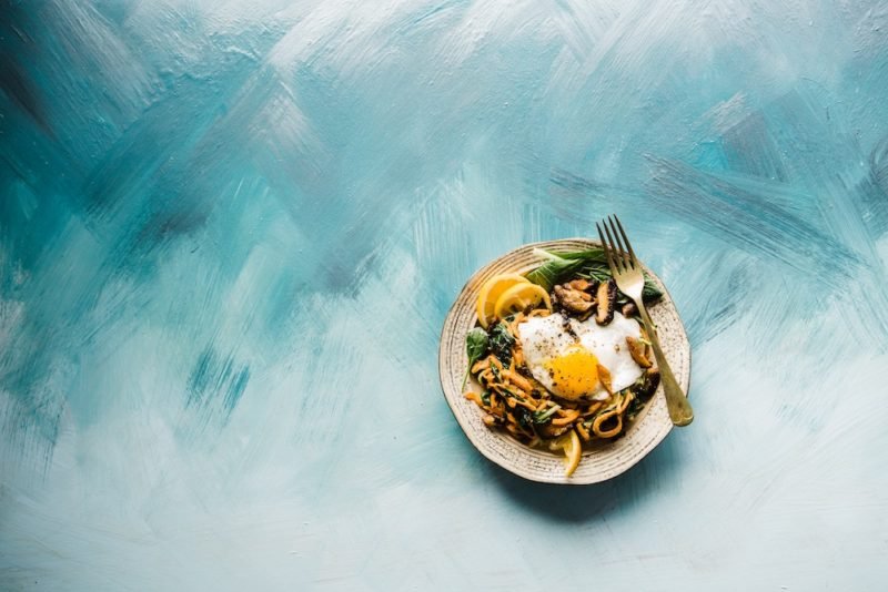 Honoring hunger means eating when your body tells you it needs food. Photo of a rustic pottery bowl of noodles, greens, lemon slices and cooked Asian mushrooms with a fried egg on top, set against a multi-hued blue background.