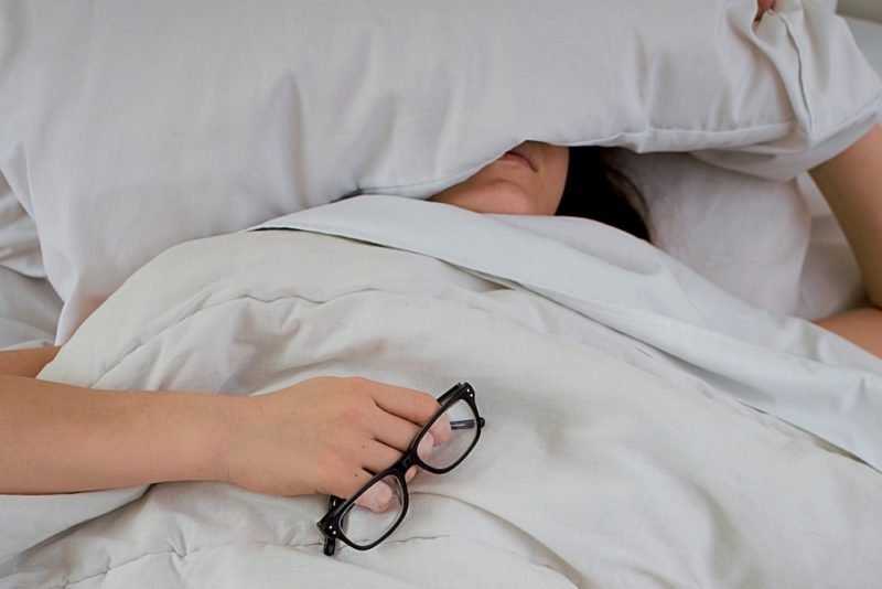 Nutrition myth busting can be hard work. Photo of a woman laying in bed under a white comforter, holding a pair of eyeglasses, with her face mostly covered by a pillow.