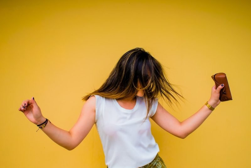 Enjoyable exercise could simply be dancing when the mood strikes you, as in this photo of a young woman with long, dark blond hair and a white sleeveless T-shirt, dancing in front of a yellow wall, holding a brown leather glasses case.