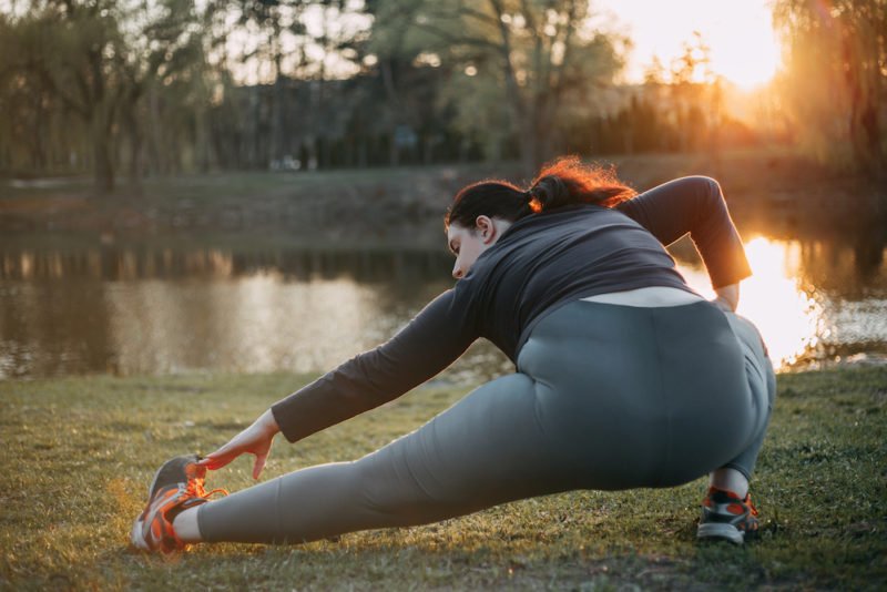 Enjoyable exercise feels good while you are doing it, as in this photo of a young woman with long, dark hair in a ponytail, and black and gray leggings and long-sleeve top, doing a deep side lunge stretch in a park.