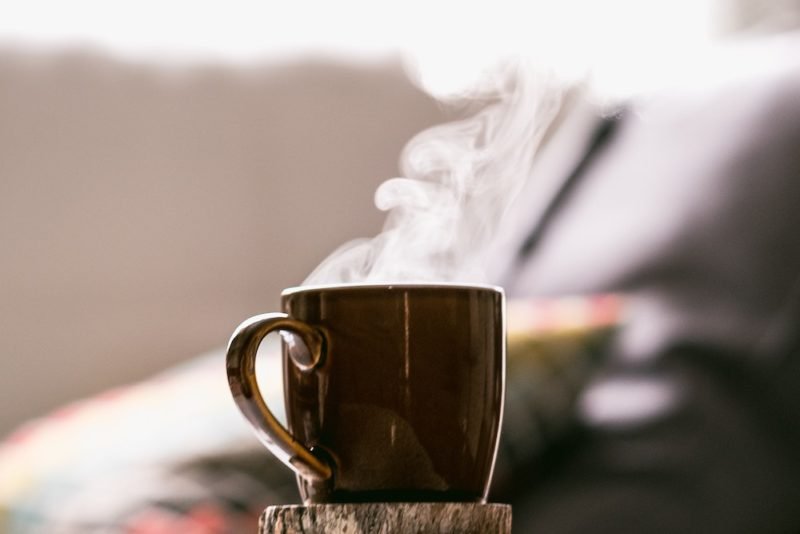 Photo of a brown mug, on a chunky wood coaster, with steam pouring out of the top. Many people consider a cup of coffee or tea part of their morning rituals.