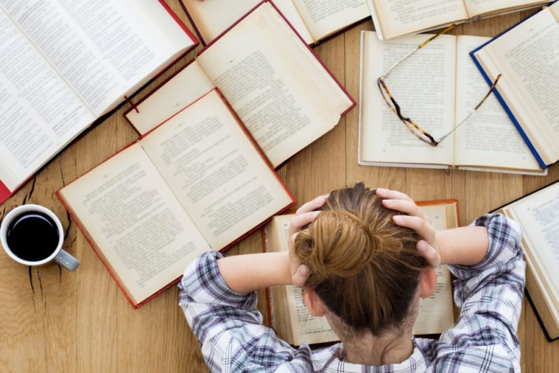 Motivation can help you achieve things, but one of the pitfalls of motivation is not knowing when to stop. Overhead photo of a young woman with dark blond hair holding her head in her hands while sitting a a table covered in textbooks and notebooks.