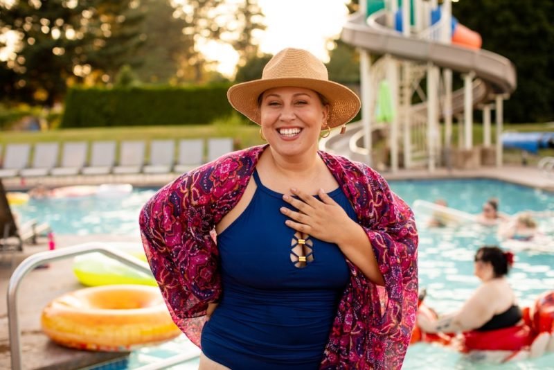 Photo of a woman in a larger body demonstrating summer body confidence by wearing a deep blue one-piece bathing suit, dark pink print sheer cover up and straw hat as she stands smiling by an outdoor pool.