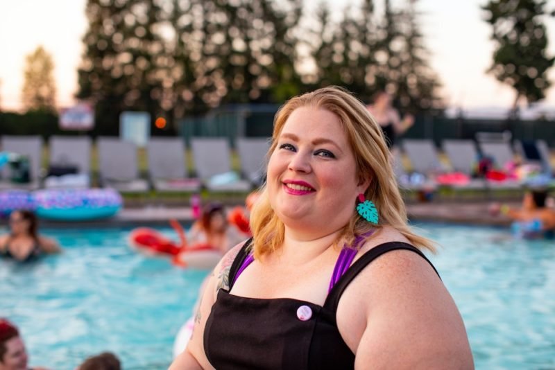 A blonde-haired woman in a larger body showing summer body confidence as she poses, smiling, in a purple halter-neck bathing suit under a black sundress in front of an outdoor pool.