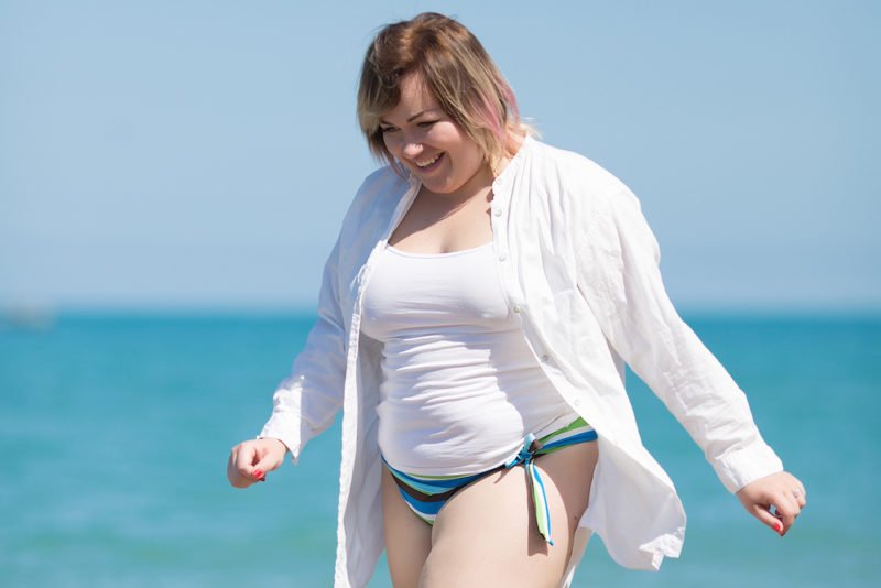 A blonde woman in a larger body showing summer body confidence as she walks on the beach, wearing green and blue-striped bikini bottoms, a white tank top and an open white long-sleeved shirt.