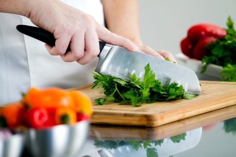 Improve health without dieting by eating nutritious food. Photo is a close up of a woman's hands chopping flat-leaf parsley on a wooden cutting board, with bowls of red, orange and yellow peppers next to her.