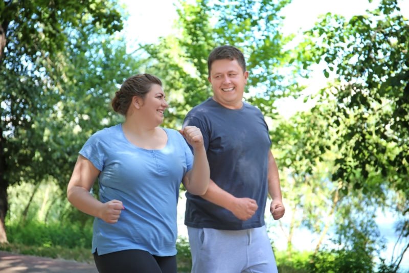 Improve health without dieting by getting regular physical activity. Photo of larger-bodied man and woman in their 30s going for a power walk, smiling, on a tree-lined road next to a pond or lake.