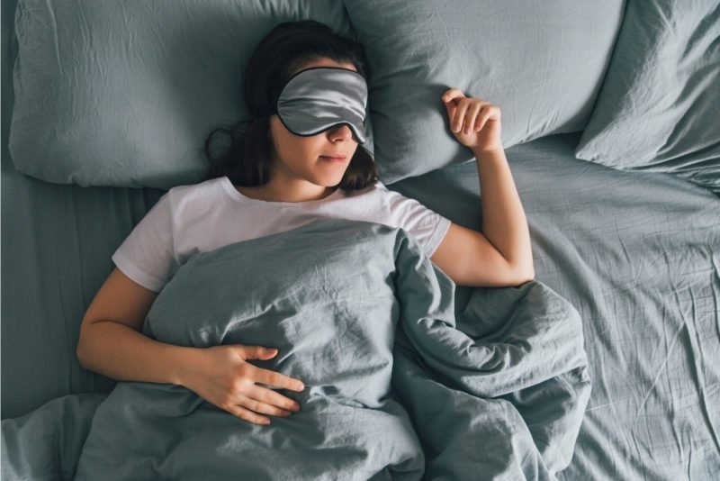Improve health without dieting by getting a good night's sleep. Photo of a young dark-haired woman in a white T-shirt and a gray silk sleep mask, sleeping on her back in a bed with gray sheets and duvet cover.