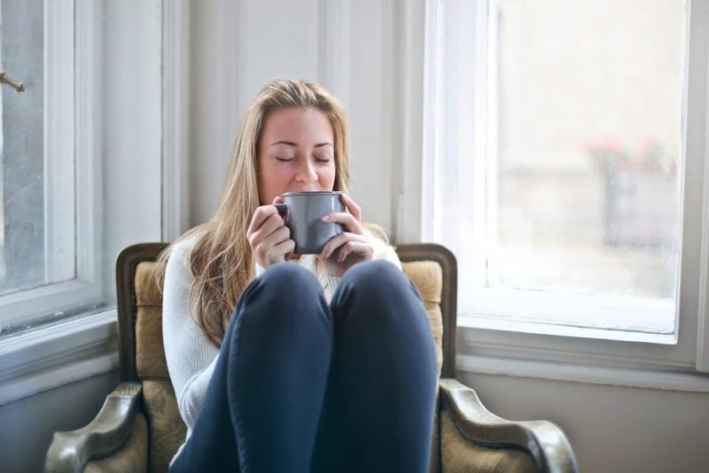 Improve health without dieting by having healthy ways to cope with stress. Photo of a woman sitting on a vintage chair in the corner of a room, flanked by windows. Her knees are drawn up, and she's enjoying a cup of tea, with her eyes closed. She's wearing jeans and a white sweater, and has long blonde hair.