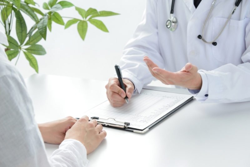 Improve health without dieting by getting regular preventive healthcare. Close up photo of a doctor and a female patient wearing a white linen shirt sitting across from each other at a desk with a houseplant while the doctor fills out a form on a clipboard.