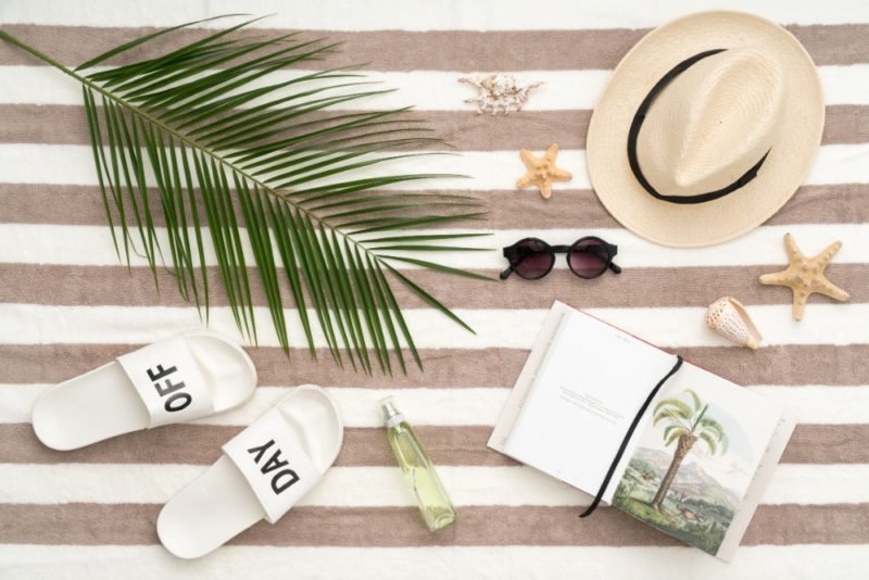 Many people don't take all of their available vacation time, missing out on some of the benefits of downtime. Photo of a tan-and-white striped beach towel with a sun hat, white slide-on shoes that say "Day" and "Off," sunglasses, seashells, a large palm frond and a vacation book arranged on top.