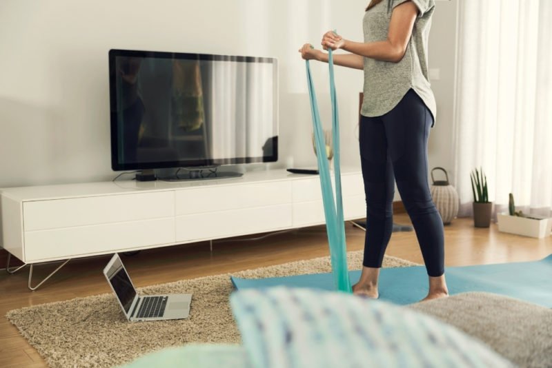 One way to improve self care in the new year is to find enjoyable forms fo movement. Photo of a young woman standing on a yoga mat in front of her laptop using a resistance band.