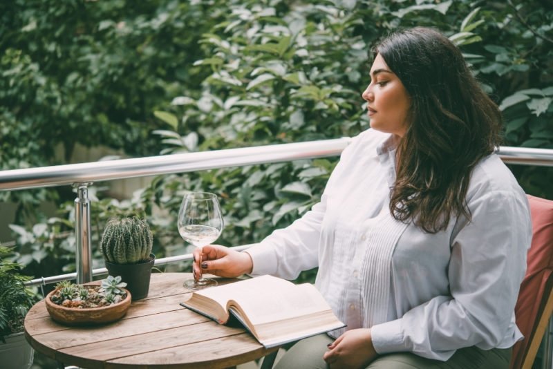 Photo of a young woman in a larger body with long dark hair and olive skin, looking contemplative, sitting at a small round wooden outdoor table near a metal deck railing, with a book open in front of her and a glass of white wine in her hand.