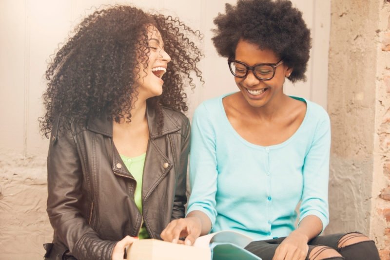 Belonging is important for health. Photo of two young black women laughing and looking at a book together.
