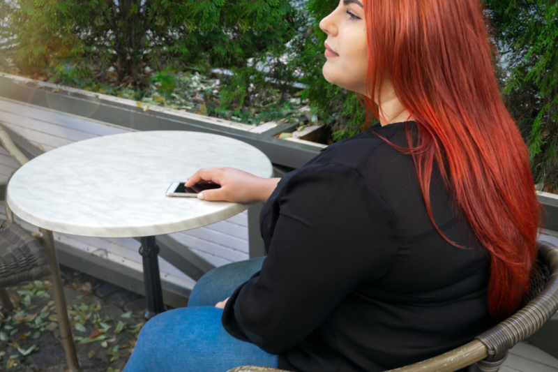 How to talk about fat bodies: Photo of a young small fat woman with long, brilliant red-orange dyed hair and olive skin, sitting at a marble-topped bistro table outdoors next to planters holding evergreen shrubs, wearing blue jeans and a long-sleeved black top, resting one hand on her phone on the table, looking like she's waiting for someone.