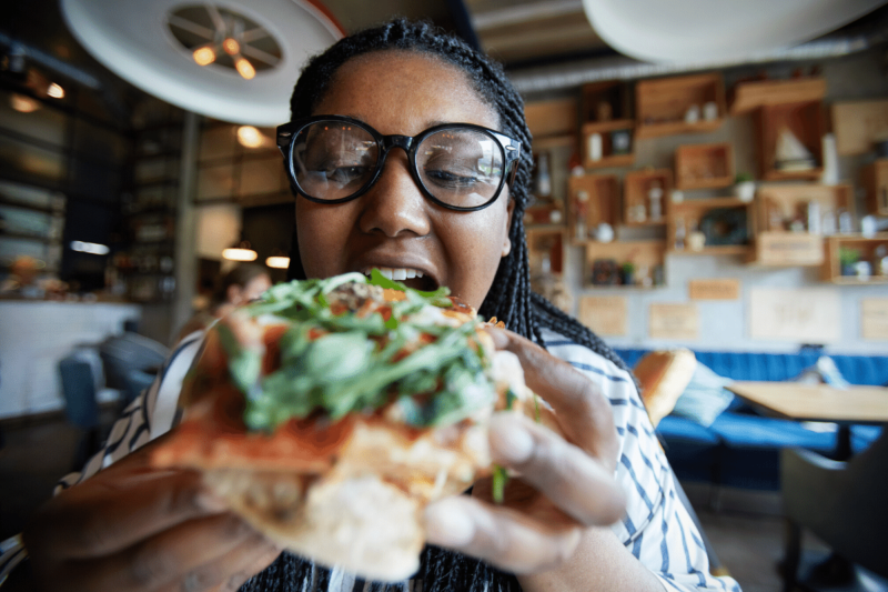 Black woman with long braided hair and big black-framed eyeglasses eating and enjoying a slice of pizza topped with arugula