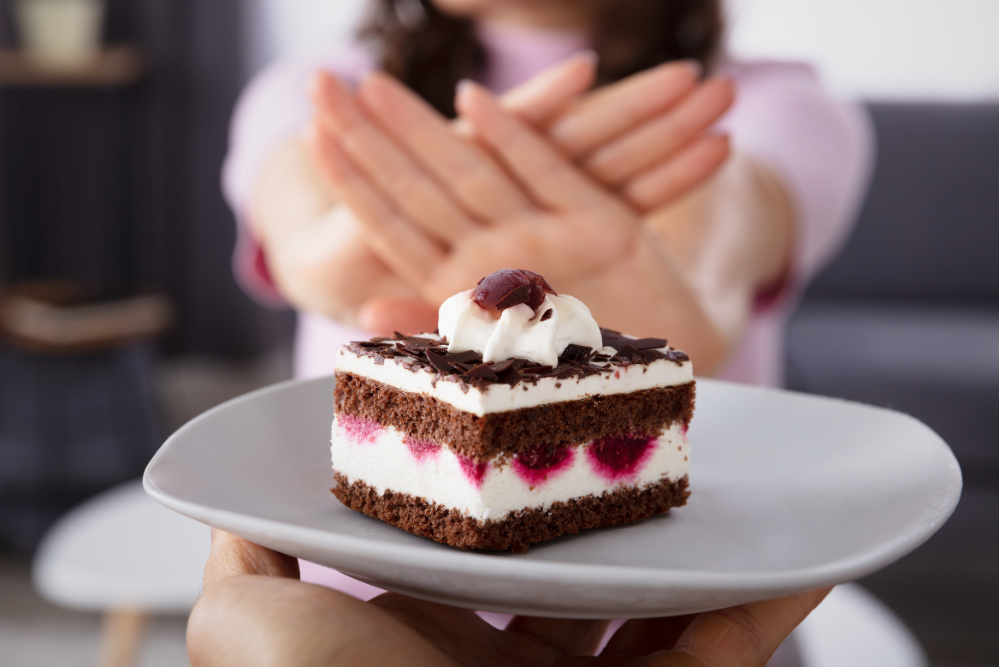 Close up of a piece of birthday cake in front of a woman's hands held up in a "no" gesture