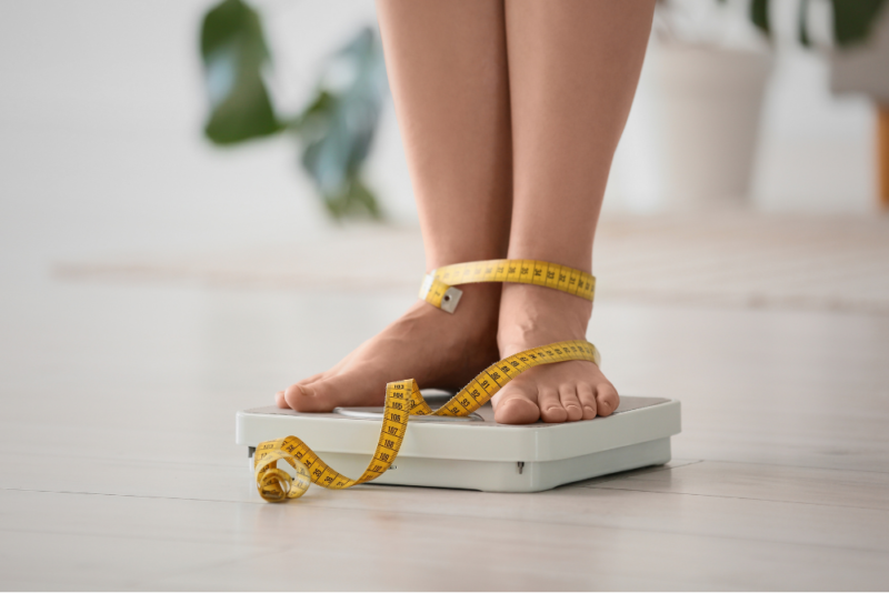 A woman's lower legs, ankles wrapped in a tape measure as she stands on a scale, emblems of self-objectification