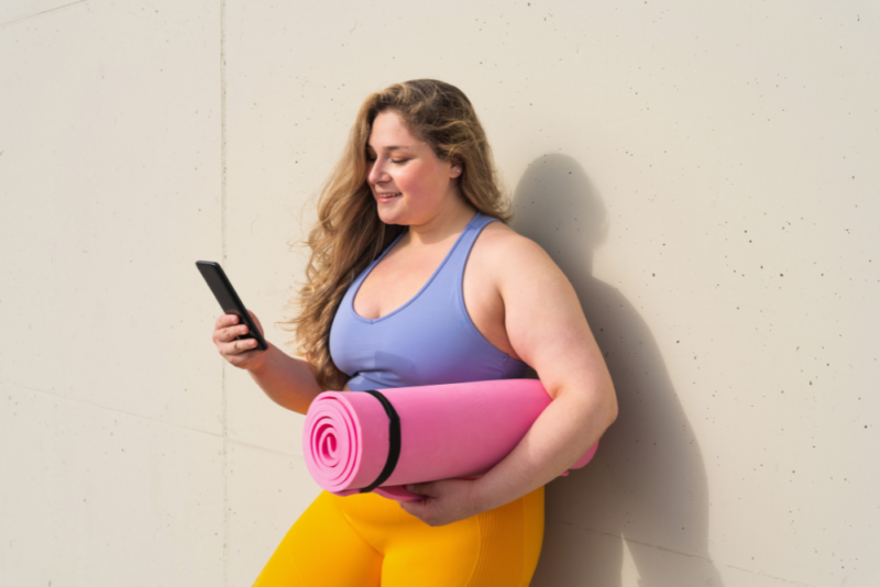 Smiling woman in a larger body looking at her phone with a rolled up yoga mat under her arm.