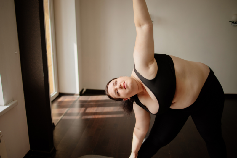 A woman in a larger body looking peaceful while doing a yoga pose while wearing black leggings and a sports bra