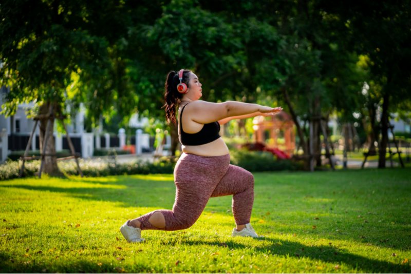 A young woman in a larger body doing a lunge in a park while wearing pink headphones, dark pink patterned leggings and a black sports bra