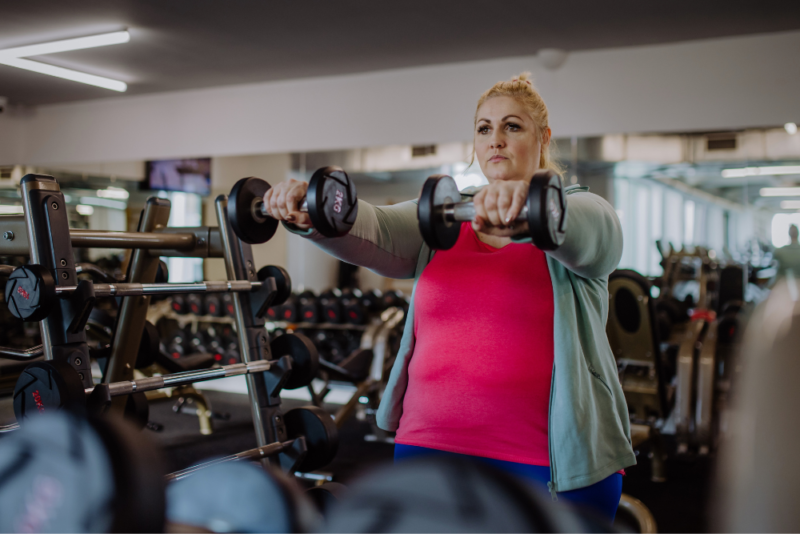 A woman in a larger body lifting free weights while wearing a bright pink top, bright blue leggings and and a light green knit workout jacket
