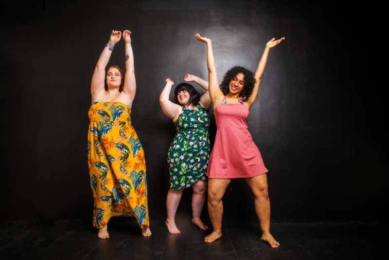 Three women in average-to-plus size bodies wearing summery dresses and dancing joyously with their hands over their heads, feeling free of internalized weight bias.